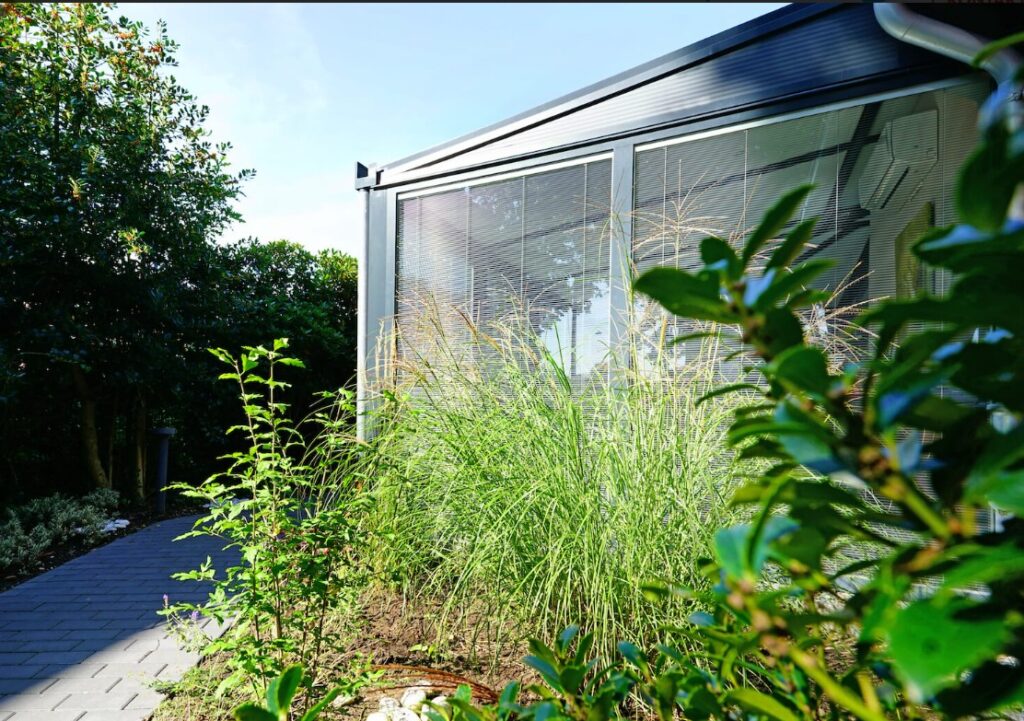 Ein Wintergarten im Sommer, der leicht verdeckt wird von der Begrünung des Gartens. Die Seiten haben große Glasflächen, der Wintergarten ist sonst aus Aluminium gebaut.