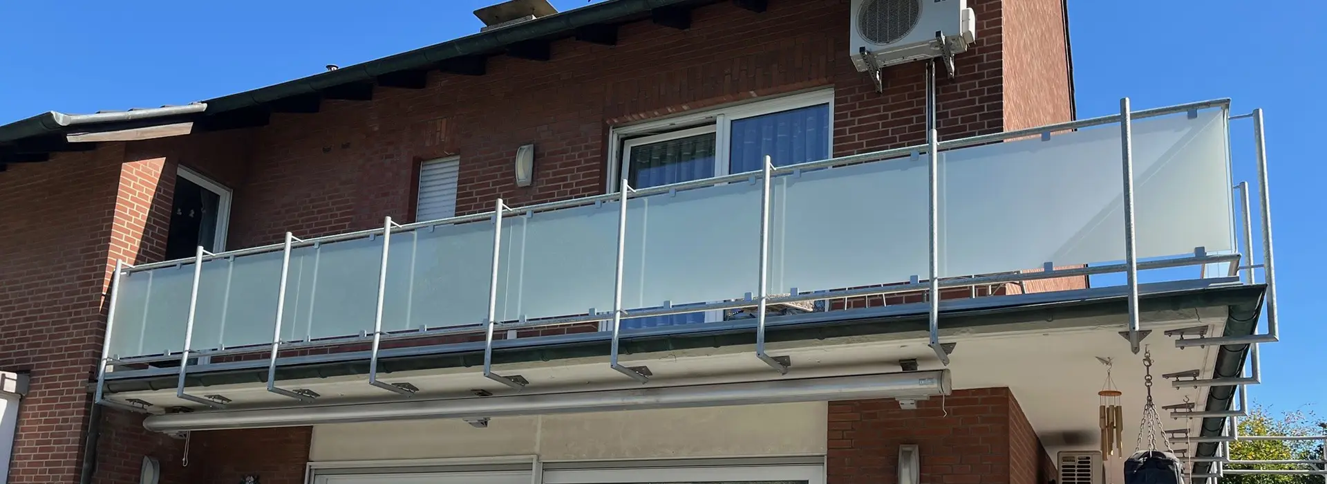 Ein Balkon im zweiten Stock mit Milchglasfenstern und Metallgeländer, der an einem Backsteingebäude angebracht ist, unter einem strahlend blauen Himmel.