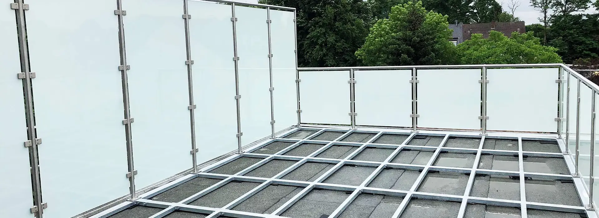 Balkon mit grauem Metallgeländer und transparenten weißen Sichtschutzplatten, umgeben von grünen Bäumen.